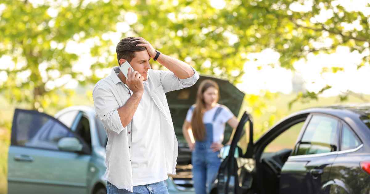 A man on the phone appearing frustrated. A woman stands behind him, and it appears two cars collided